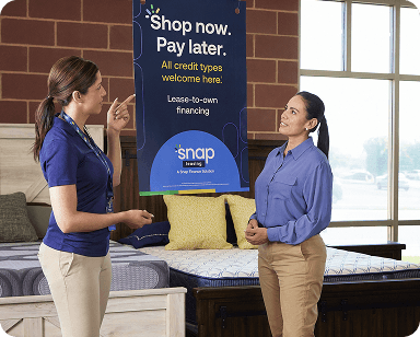 Two people in a store discussing a "Shop now. Pay later." sign for lease-to-own financing by Snap above a display bed.