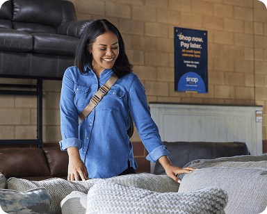 A woman in a denim shirt smiles while touching a sofa in a furniture store. A sign in the background reads, "Shop now. Pay later."