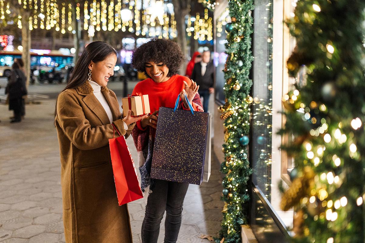 Two women smile while shopping at night, holding gift bags and a wrapped present near a festively decorated store window.