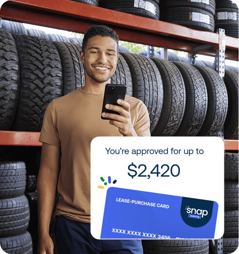 Man holding a phone in front of a rack of tires, smiling at a lease-purchase card approval notification for up to $2,420.
