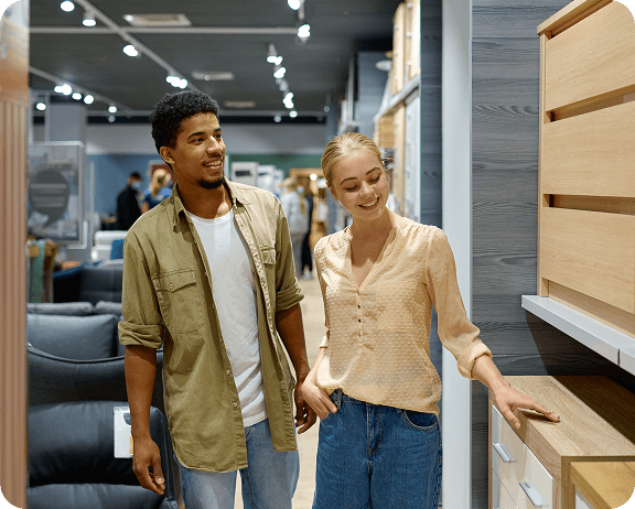 A smiling couple examines modern wooden cabinets in a furniture store, surrounded by various home furnishings.