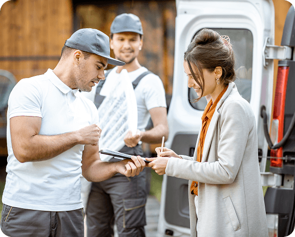 Woman signs on a clipboard held by a delivery worker next to a van, while another worker carries a package.