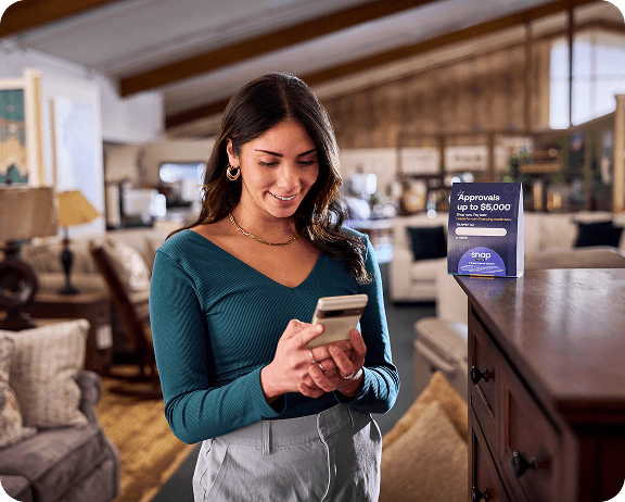 Woman using smartphone in a furniture store, standing near a sign about approval up to $5,000.