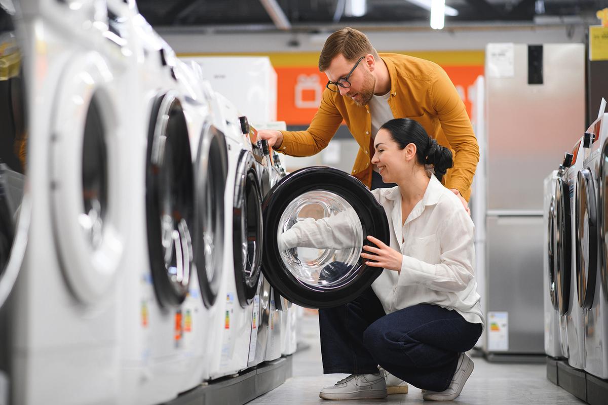 A couple inspects a front-loading washing machine in an appliance store; the woman is kneeling and opening the door while the man looks on.