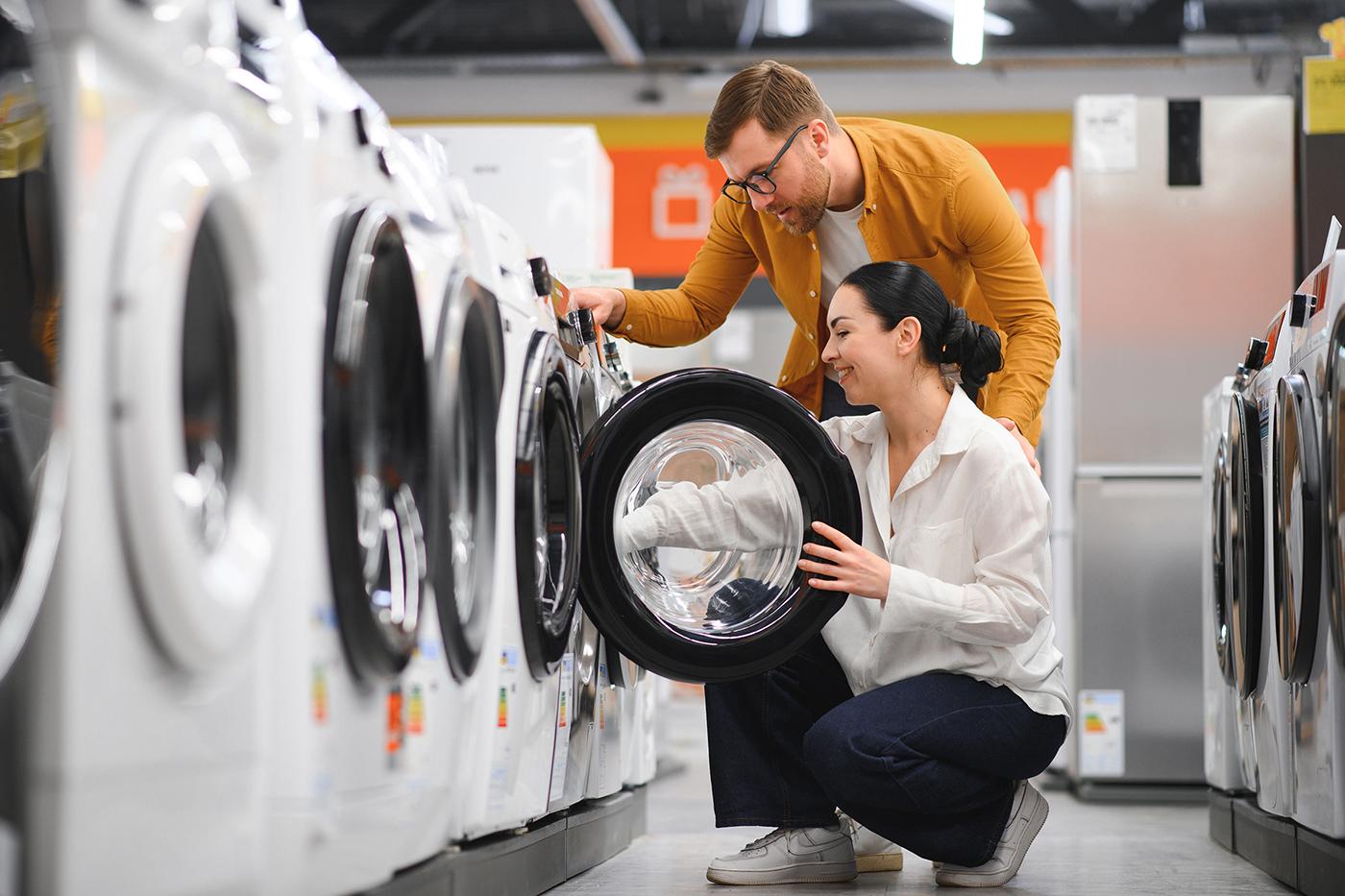 A couple inspects a front-loading washing machine in an appliance store; the woman is kneeling and opening the door while the man looks on.