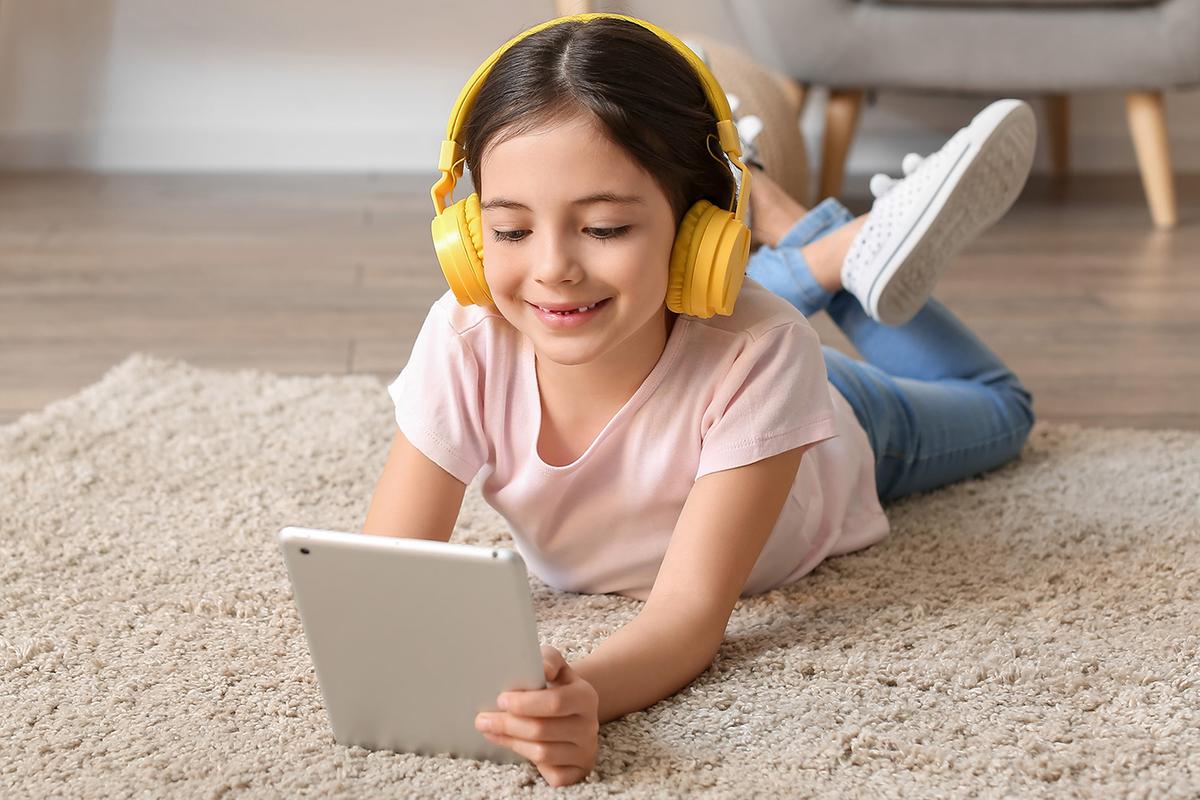 A young girl lies on a carpeted floor wearing yellow headphones and smiling while looking at a tablet in her hands.