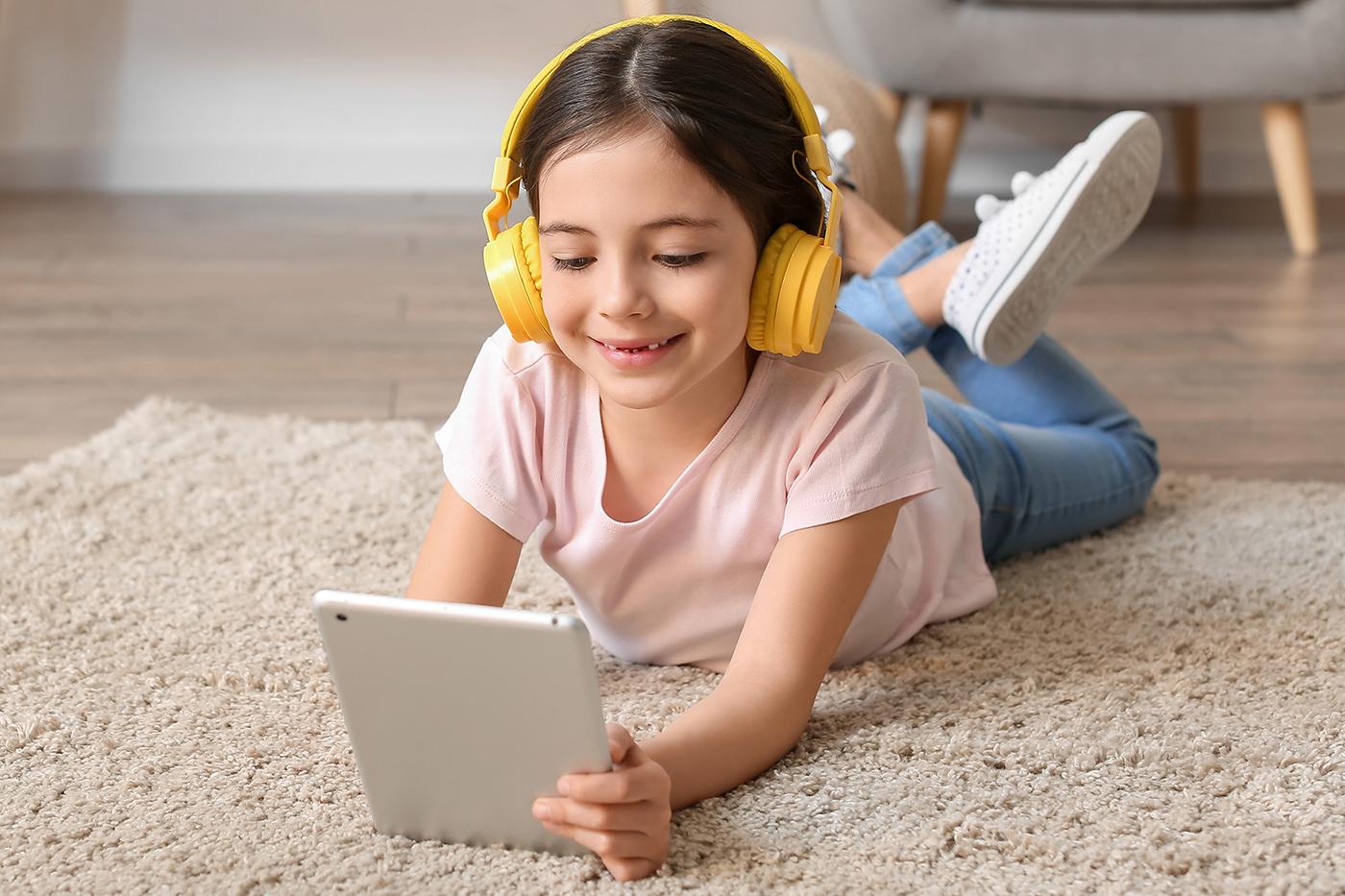 A young girl lies on a carpeted floor wearing yellow headphones and smiling while looking at a tablet in her hands.
