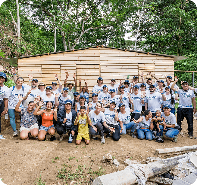A large group of volunteers wearing matching "snap" T-shirts pose happily in front of a wooden structure, surrounded by lush greenery.