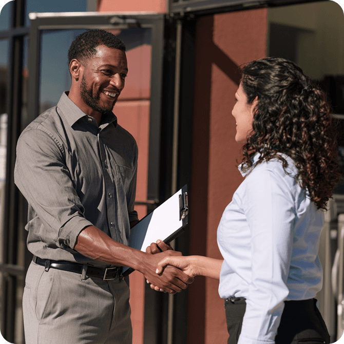 A man and woman smiling and shaking hands outside a building. The man holds a clipboard.