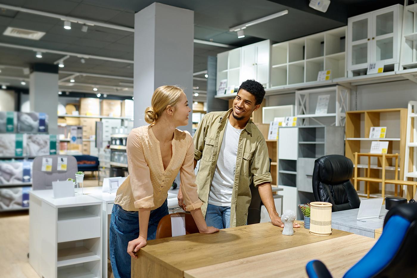 Smiling man and woman lean on wooden office desk in modern furniture store, surrounded by shelving displays and decor.