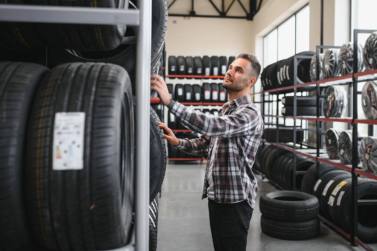 A man in a plaid shirt examines tires on a shelf inside a well-lit tire shop with rows of tires and wheels around him.