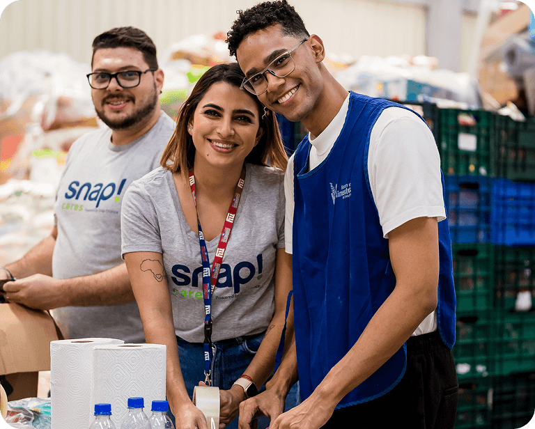 Three volunteers smiling while sorting supplies at a distribution center, wearing casual and uniform shirts with visible logos.
