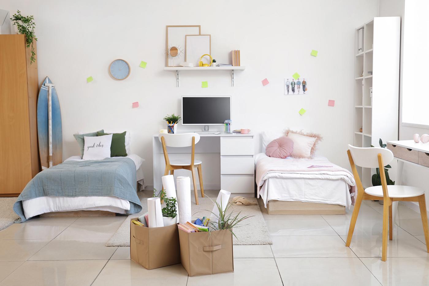 Bright twin-bed dorm room with central desk, computer, surfboard, shelving and storage bins on glossy tiled floor.