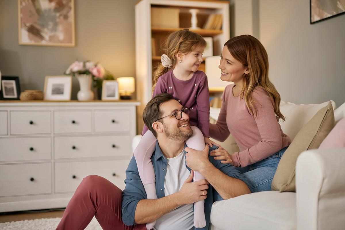 A smiling family of three sits together in a cozy living room; the father has a young girl on his shoulders while the mother sits beside them.