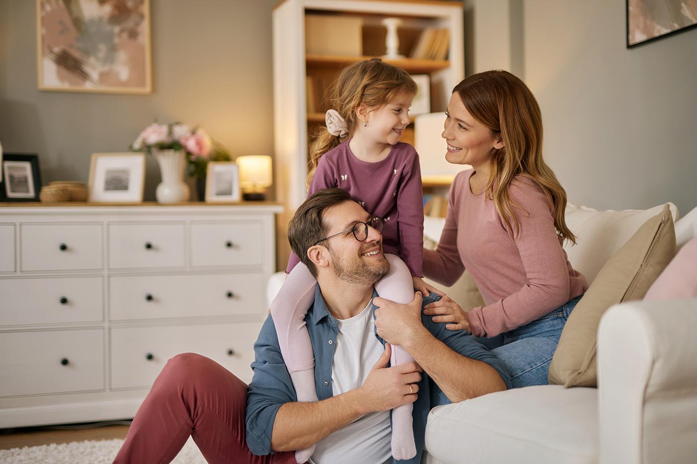 A smiling family of three sits together in a cozy living room; the father has a young girl on his shoulders while the mother sits beside them.