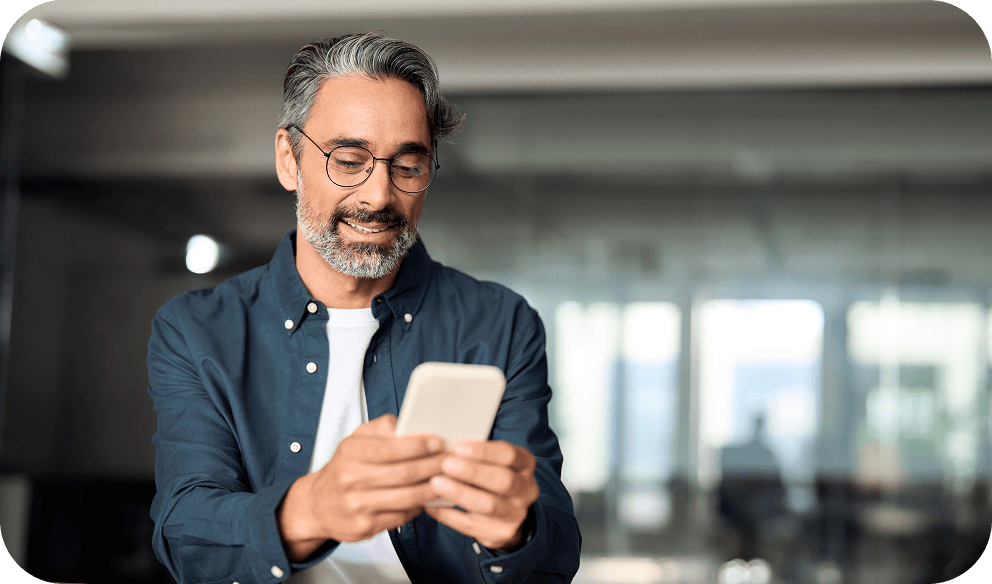A man with glasses and gray hair smiles while looking at his phone in a modern, bright office setting.