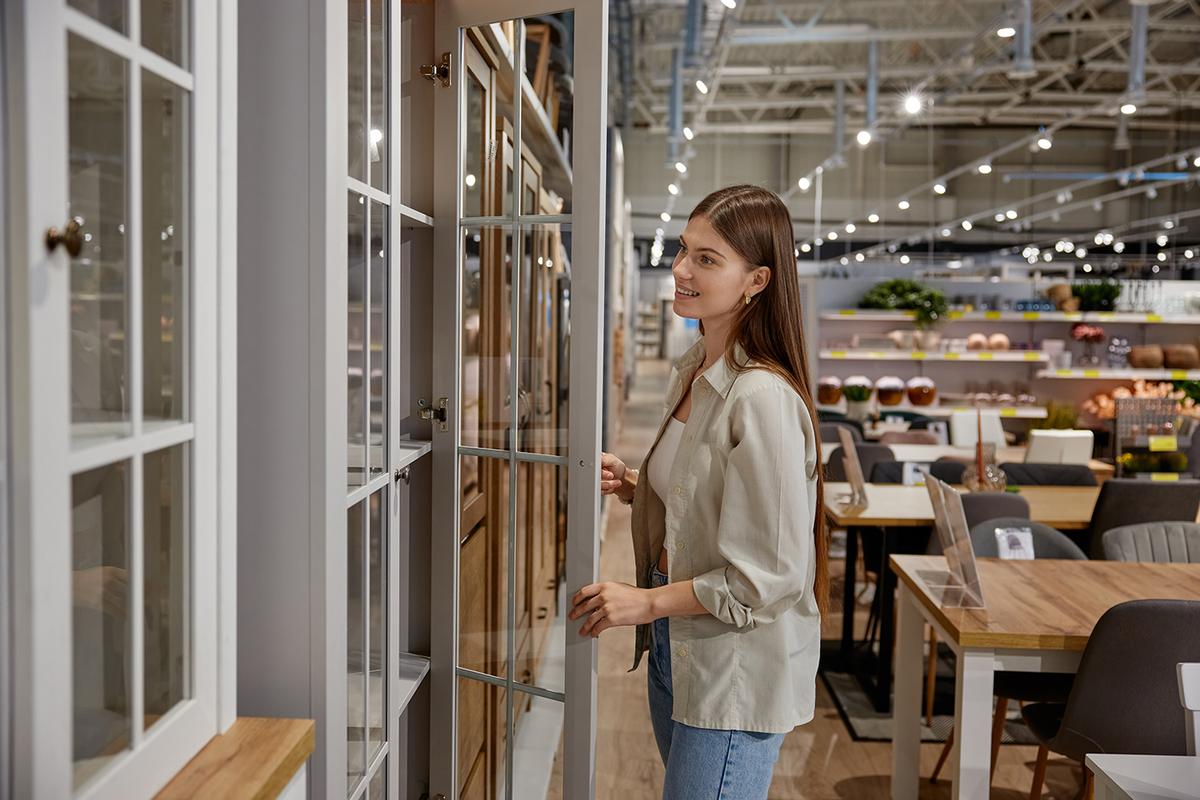 A woman in a furniture store opens the glass door of a display cabinet while browsing among tables and home decor items.