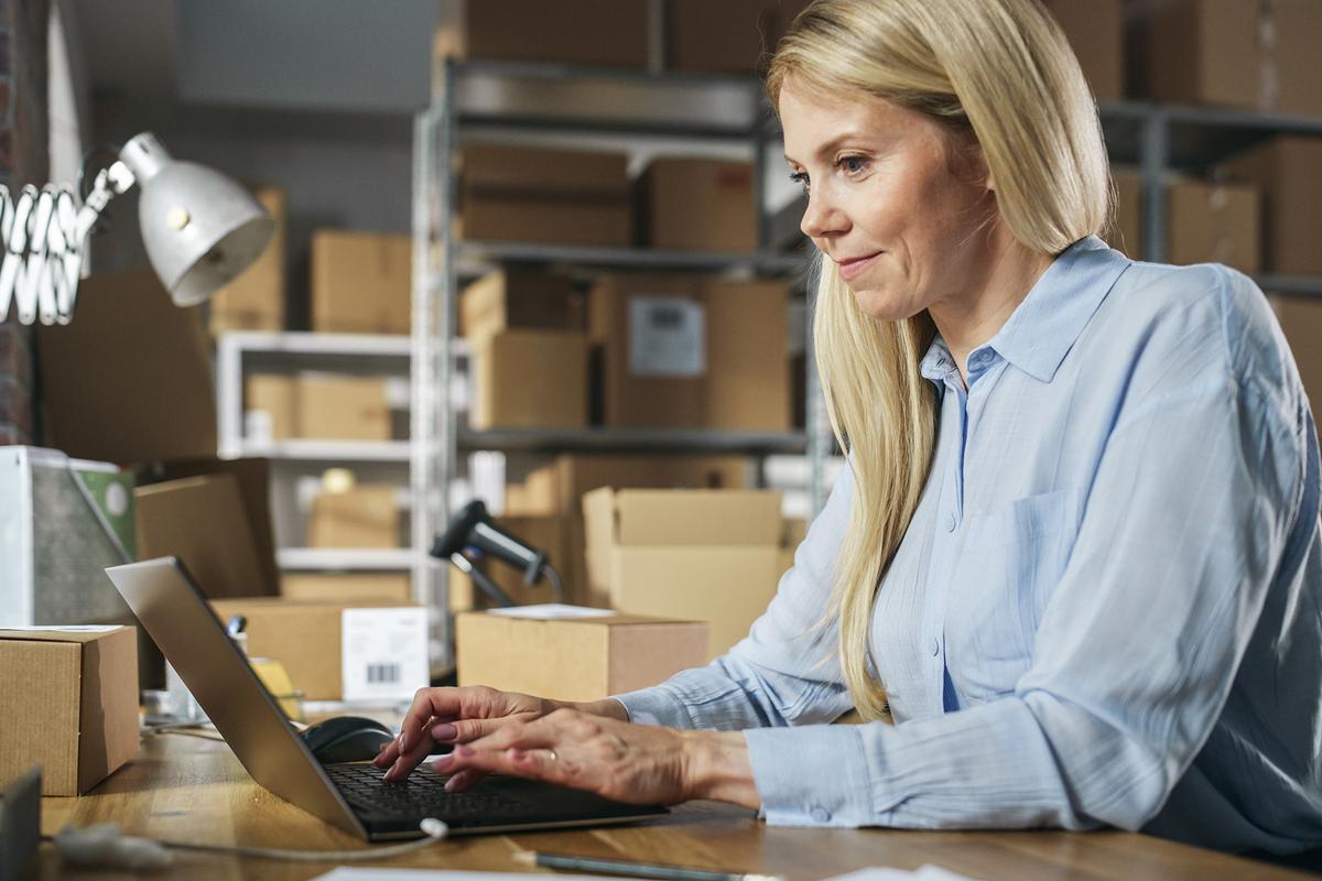 A woman works on a laptop in a warehouse setting, surrounded by shelves of cardboard boxes.