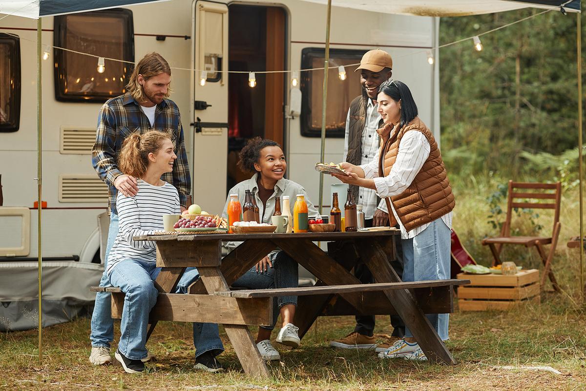 Group of five friends around a wooden picnic table outside an RV, enjoying food and drinks beneath café-style string lights.