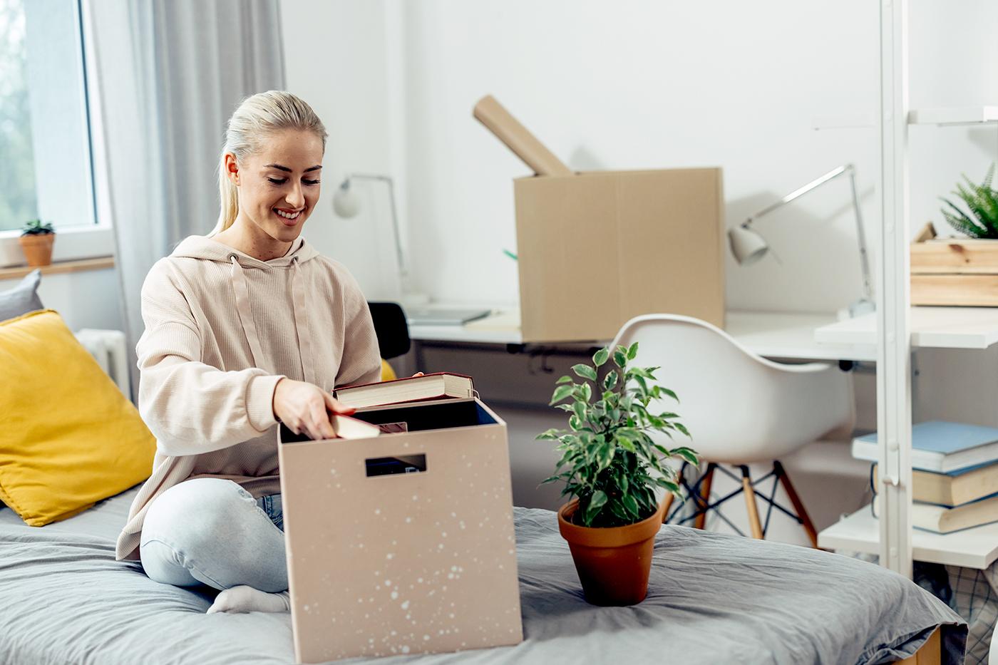 Young woman in hoodie sits on bed unpacking books from a speckled box; potted plant and moving boxes nearby.