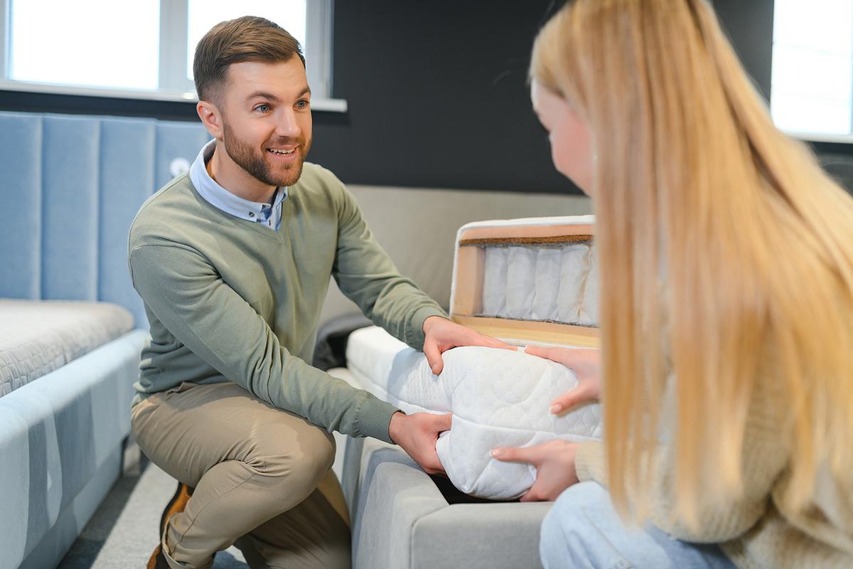 A man shows a woman a cross-section of a mattress in a store, highlighting its inner layers while they both hold it and discuss.
