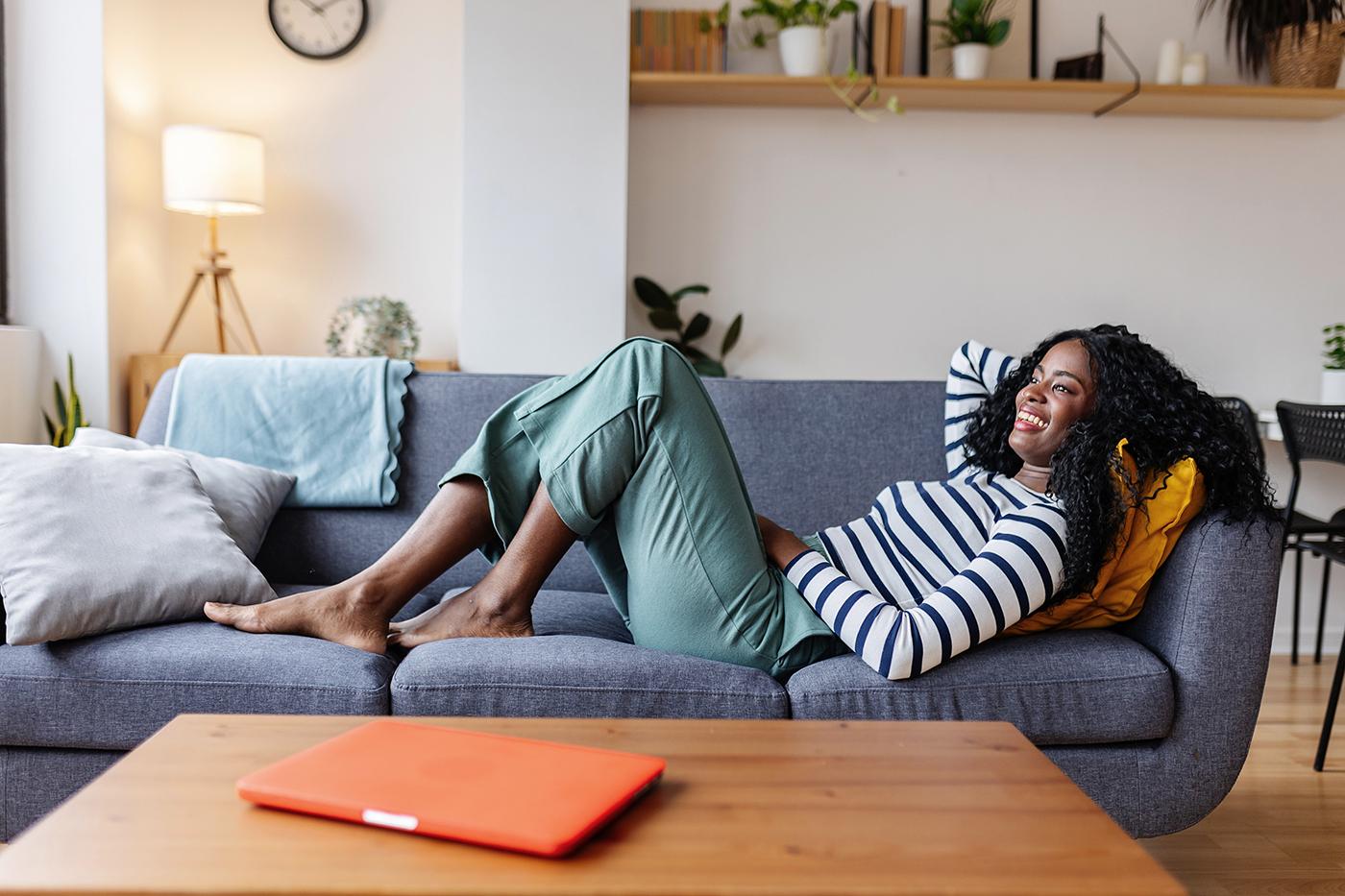 Woman in striped shirt smiles, lounging on gray sofa with legs outstretched; plants and orange laptop on coffee table.