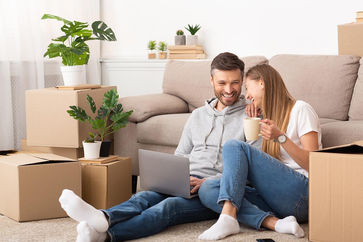 Laughing couple sit on carpet among cardboard boxes, laptop and coffee mug in hand as they settle into new home.
