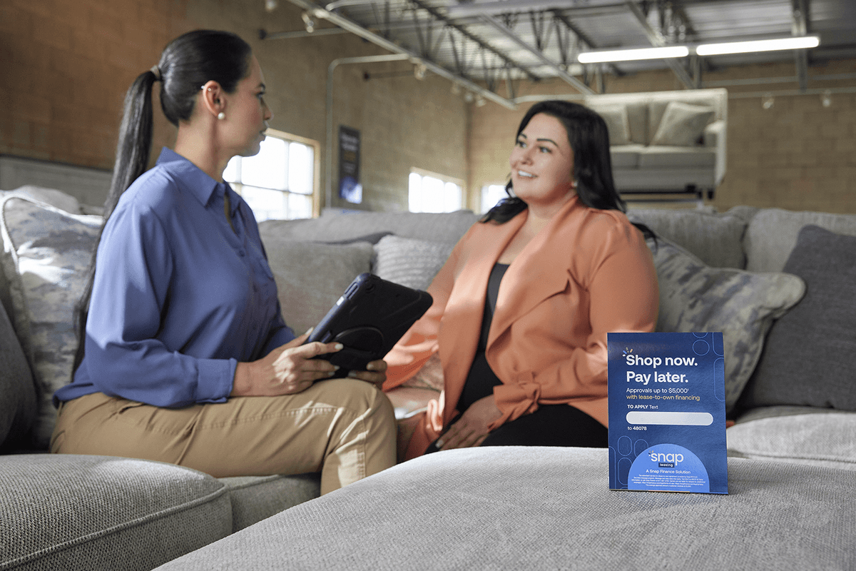 Two women talk on a sectional sofa in a showroom—one holding a tablet—while a blue sign in front reads “Shop now. Pay later.”
