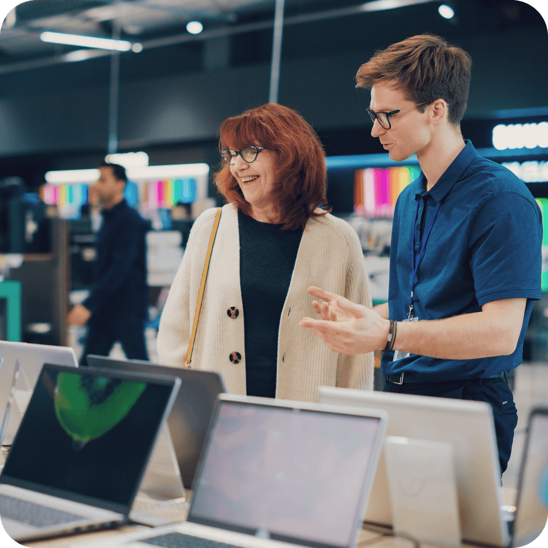 A salesperson in a blue shirt assists a smiling woman in choosing a laptop in an electronics store.