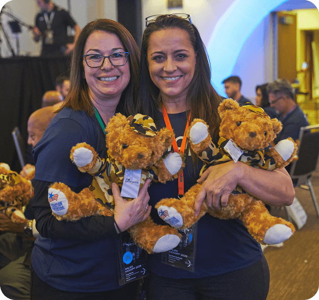 Two women smiling, each holding a teddy bear in camouflage clothing at an indoor event with people in the background.