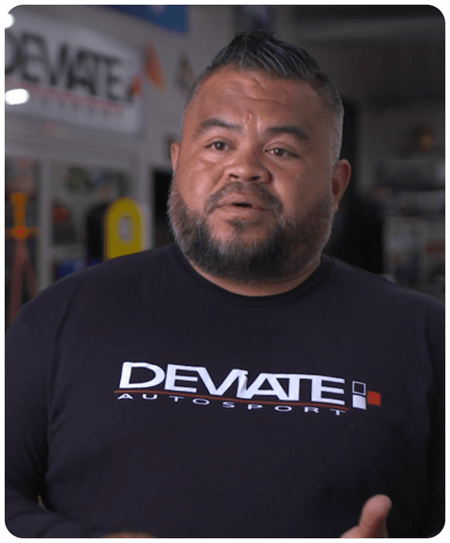 Man with a beard and short hair wearing a "DEVIATE AUTOSPORT" shirt, speaking in a workshop setting.